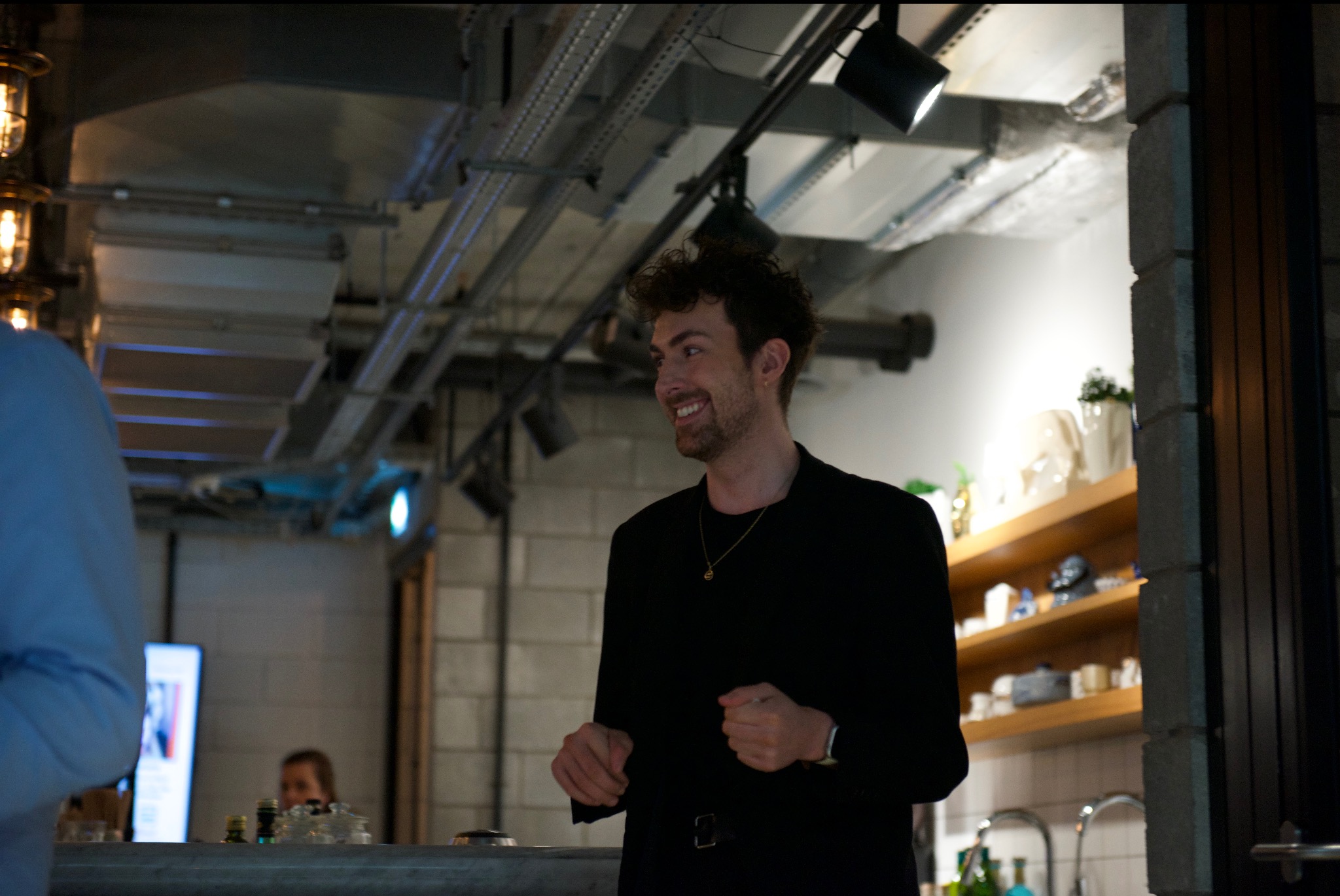 Uśmiechnięty mężczyzna w czarnej marynarce, z krótkimi kręconymi włosami i złotym łańcuszkiem, w nowoczesnym wnętrzu z widocznymi instalacjami sufitowymi i półkami z dekoracjami.