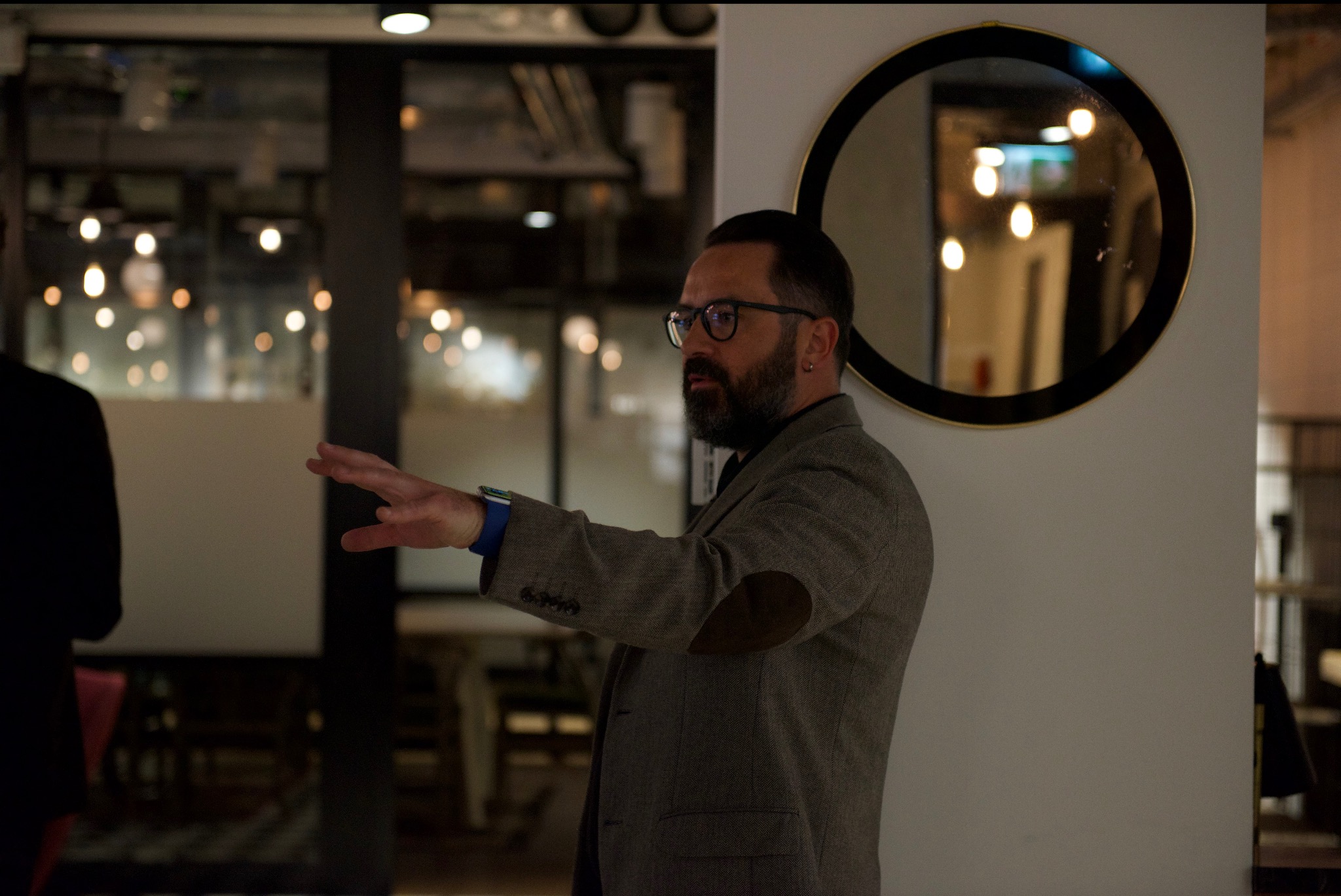 Mężczyzna w tweedowej marynarce gestykuluje w nowoczesnym biurze, obok okrągłego lustra z czarną ramą.