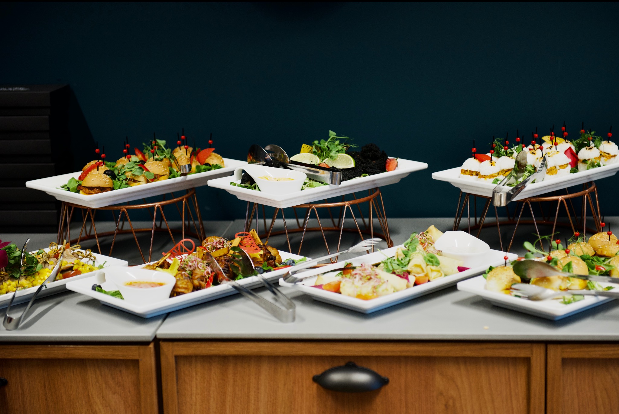 Catering eventowy: różnorodne przekąski serwowane na białych tacach na metalowych stojakach, w tym mini burgery z dekoracyjnymi szpilkami, szaszłyki i czarne danie z limonkami, szczypce do nakładania.
