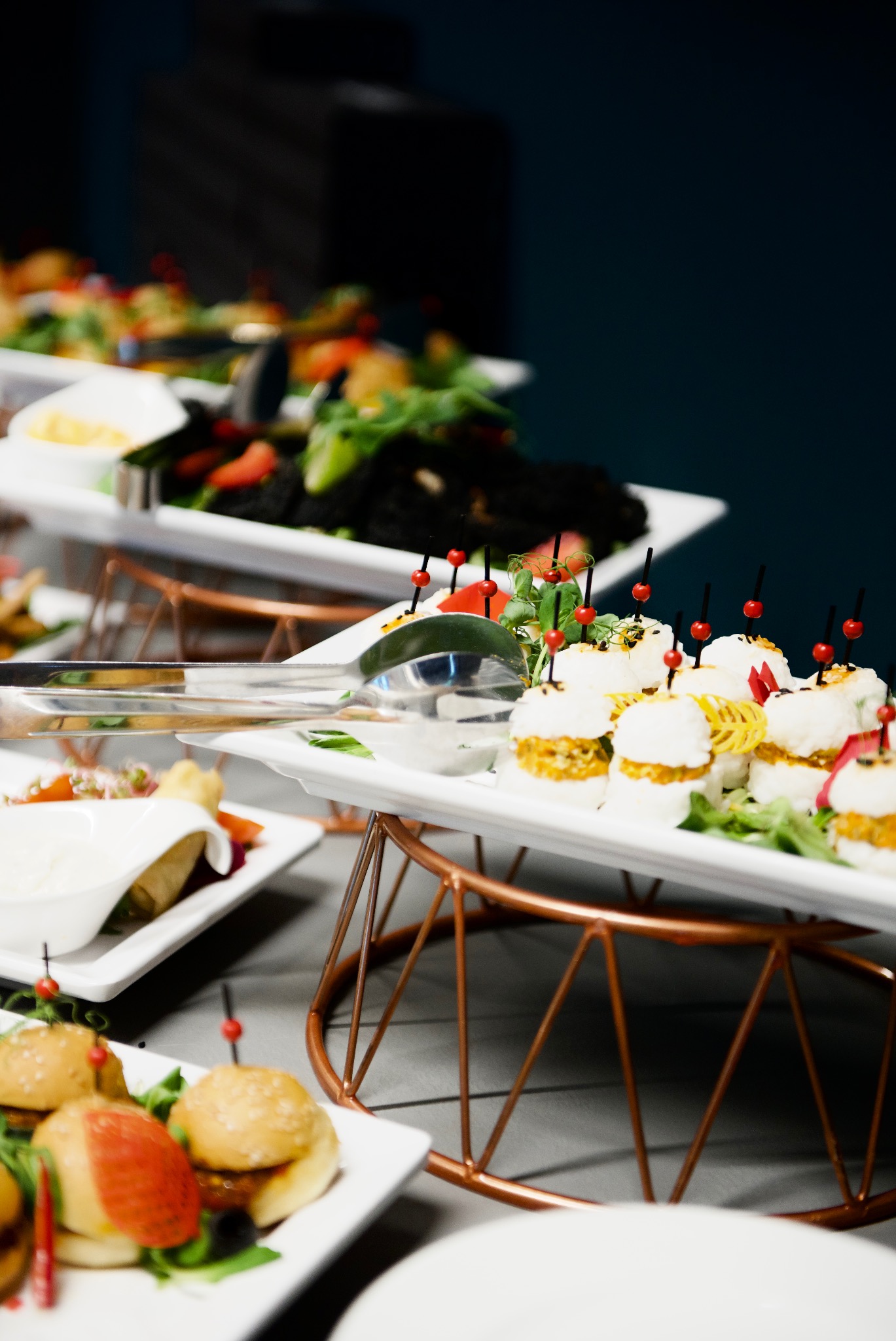 Elegancko zaaranżowany bufet z mini burgerami, przekąskami i sałatkami na białych talerzach z metalowymi stojakami w kolorze miedzi, przygotowany na przyjęcie.