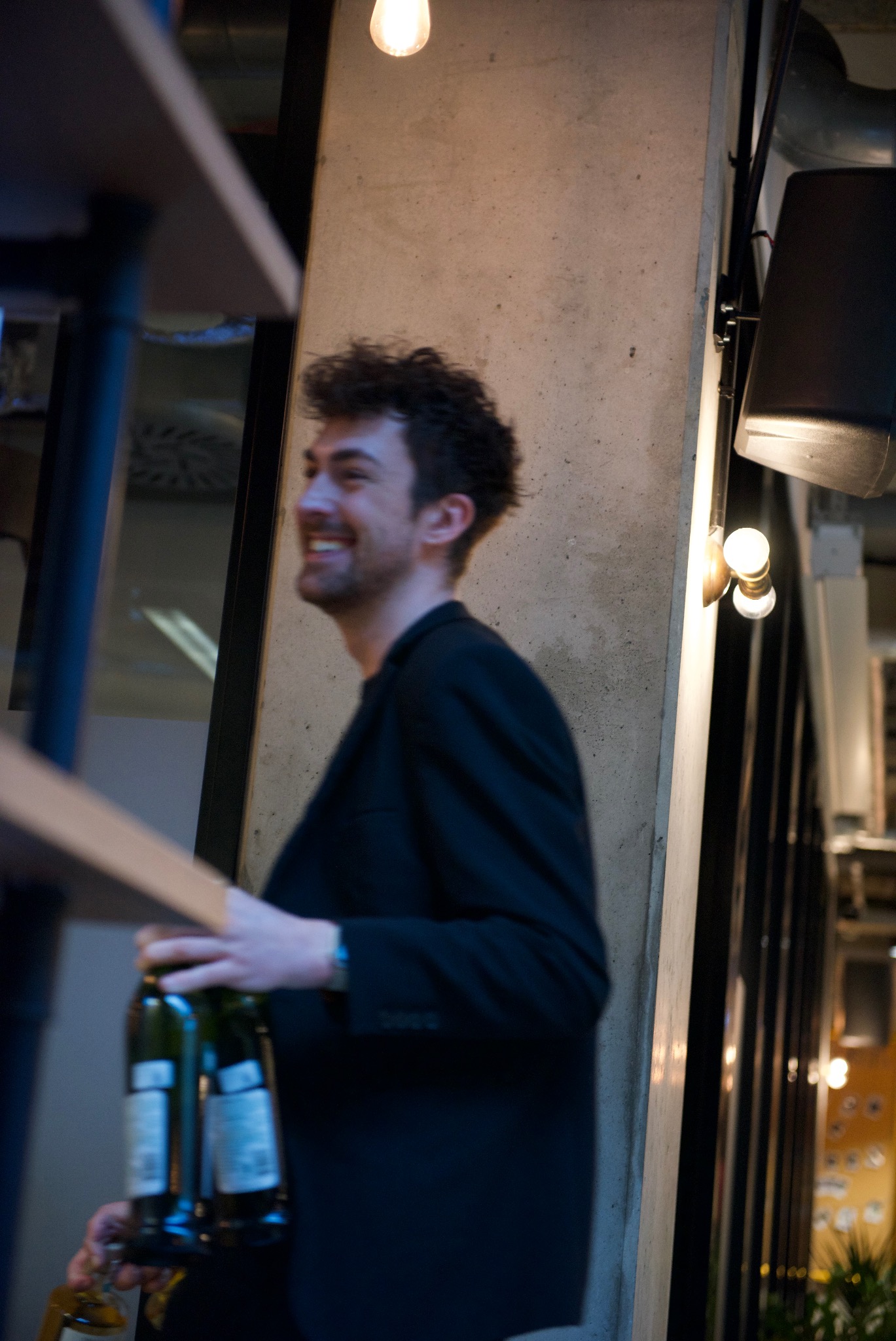 Uśmiechnięty mężczyzna w czarnej marynarce niesie butelki wina w nowoczesnym wnętrzu z betonową kolumną i dekoracyjnym oświetleniem.