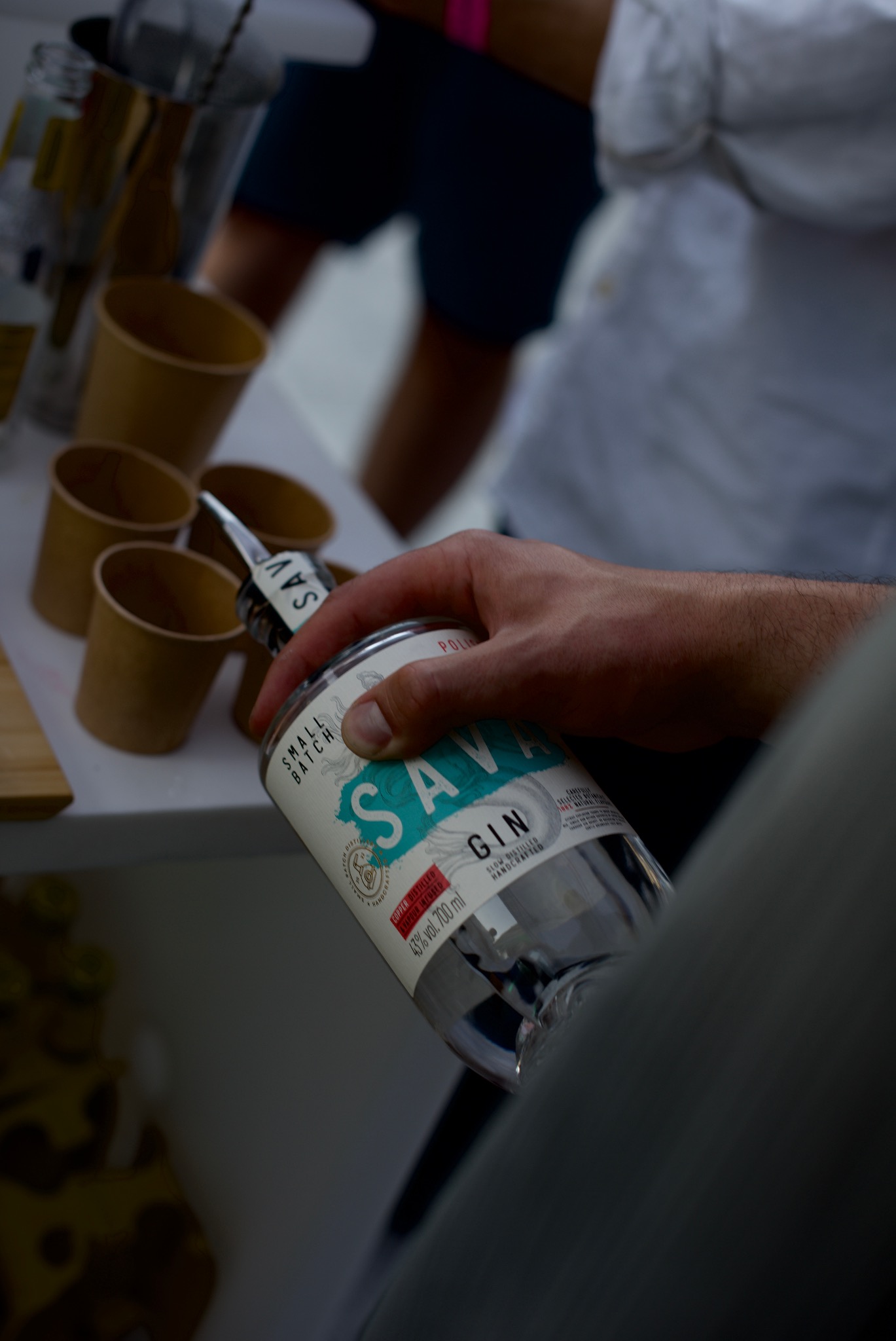 Nalewanie ginu 'Sava' z butelki do kieliszków podczas eventu, widoczne ręce osoby obsługującej i fragmenty otoczenia.