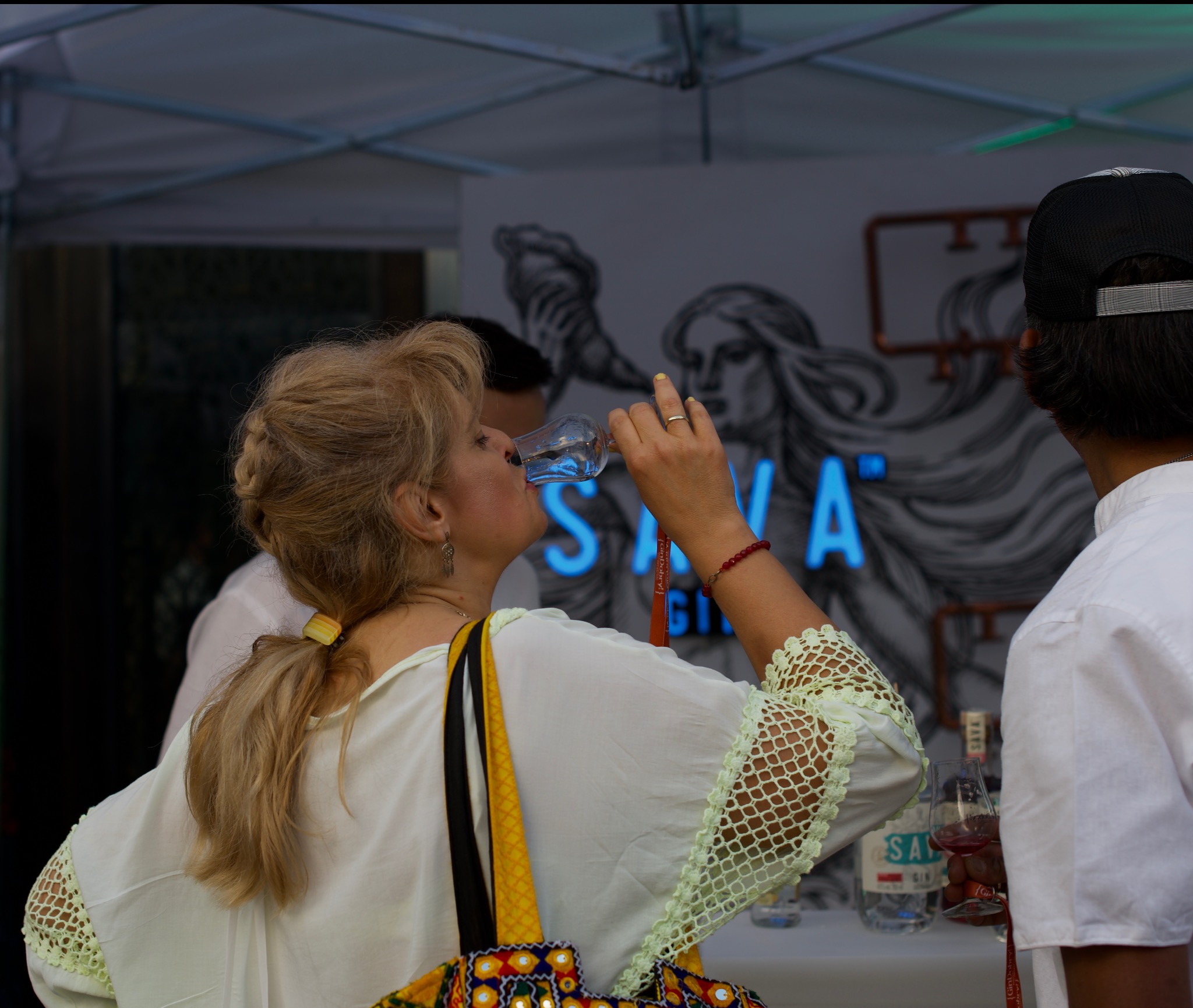 Kobieta pije gin z kieliszka na tle stoiska promocyjnego Sava Gin z neonowym logo i grafiką kobiety