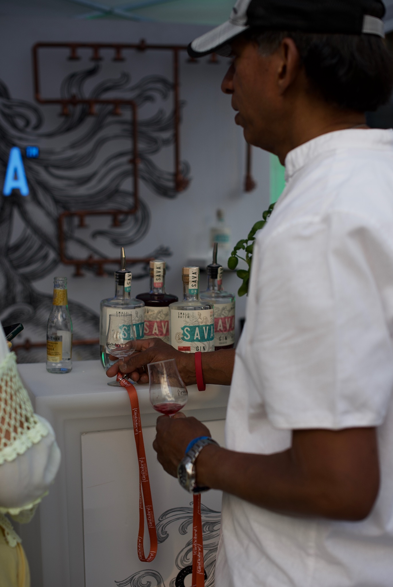 Degustacja ginu 'SAVA' podczas eventu: mężczyzna w białej koszuli trzyma kieliszek z trunkiem na tle baru z butelkami ginu i dekoracyjną ścianą.