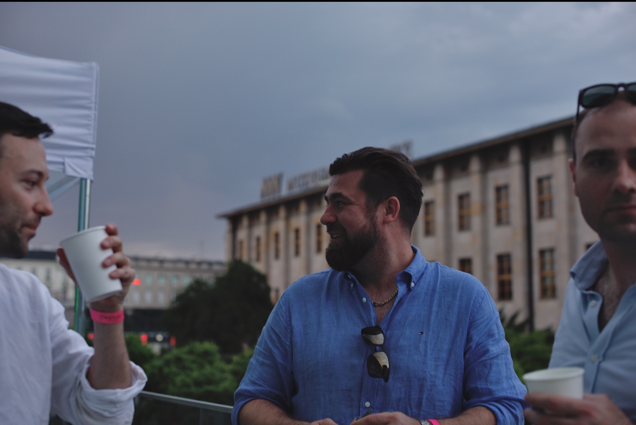 Mężczyźni w niebieskich koszulach rozmawiają podczas wydarzenia plenerowego, w tle budynek i pochmurne niebo.