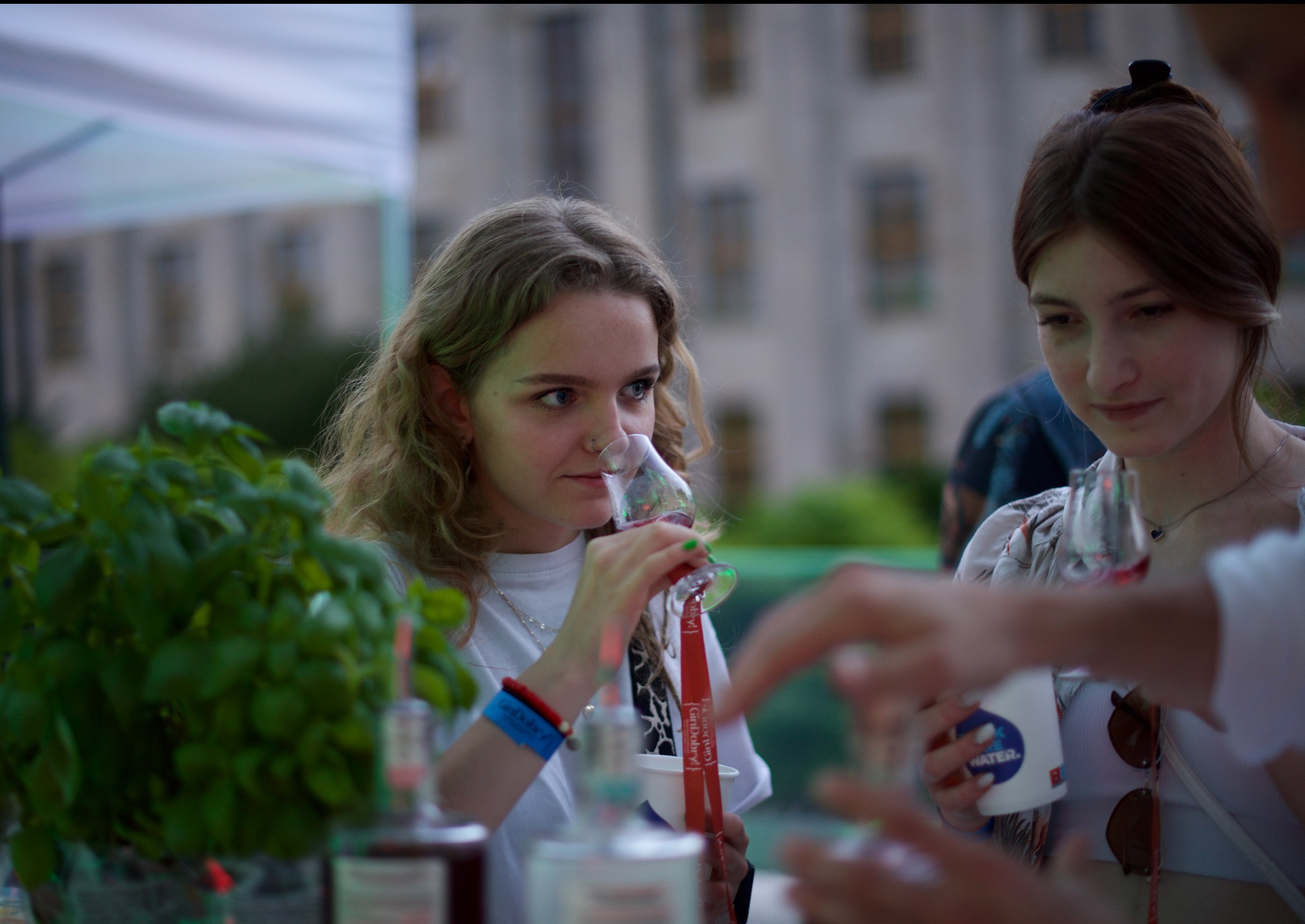 Dwie młode kobiety na imprezie plenerowej, jedna pije drinka z kieliszka na nóżce, druga trzyma kubek z napojem, w tle budynek i zieleń.