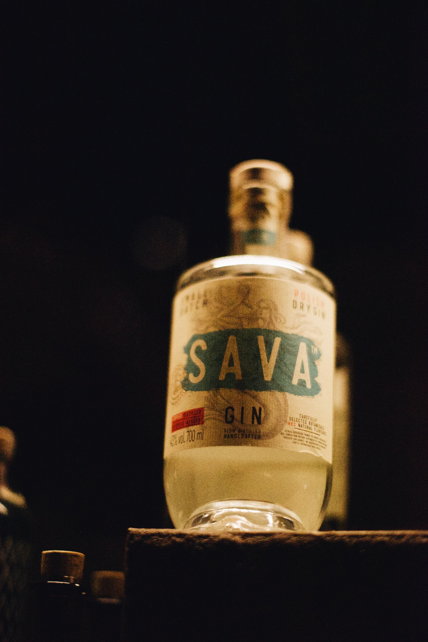 Butelka polskiego ginu Sava Dry Gin na ciemnym tle, z widocznymi detalami etykiety i innymi butelkami w tle.