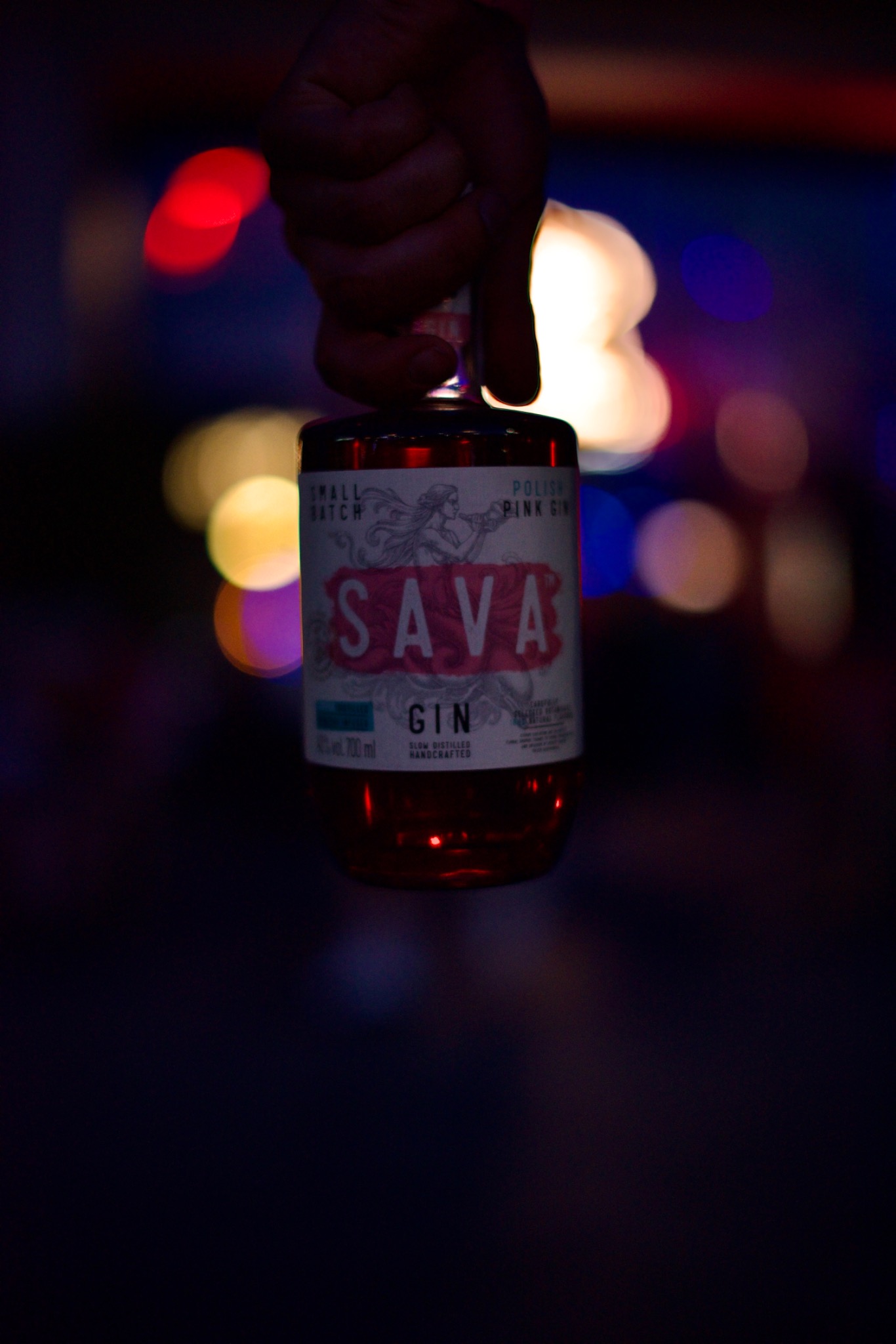 Ręka trzyma butelkę różowego ginu Sava na rozmytym tle z kolorowymi światłami bokeh.