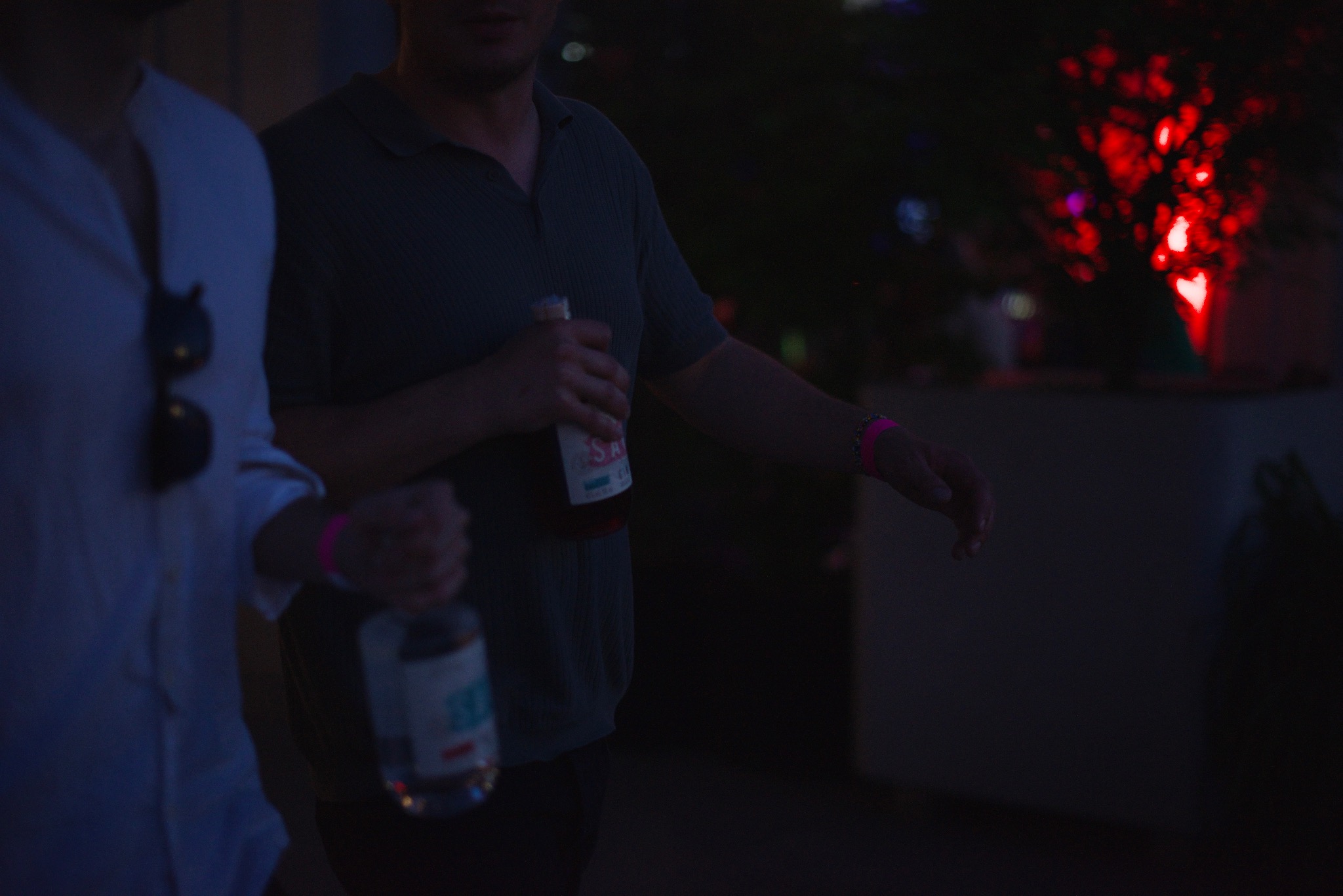 Dwóch mężczyzn trzyma butelki napojów na imprezie plenerowej z czerwonymi światłami w tle.