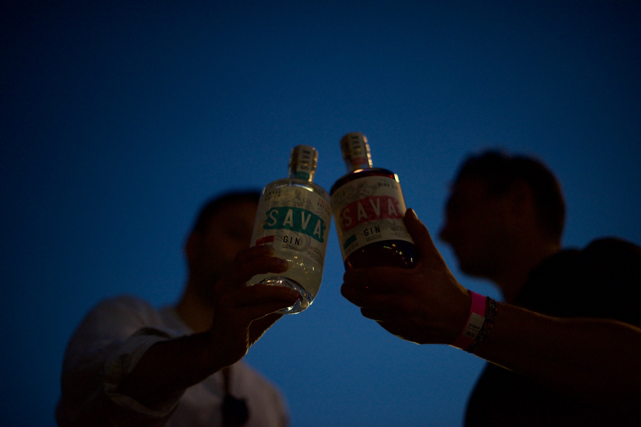 Dwie osoby wznoszą toast butelkami ginu 'Sava' na tle ciemnego, błękitnego nieba, widoczne bransoletki identyfikacyjne na nadgarstkach.
