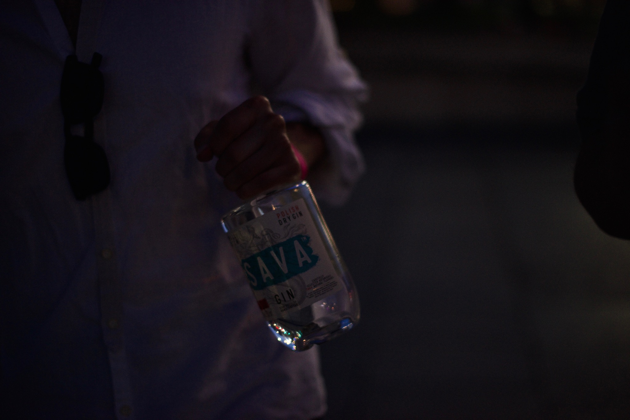 Mężczyzna w białej koszuli trzyma pod kątem butelkę ginu Sava z napisem 'Polish Dry Gin' w słabym oświetleniu.
