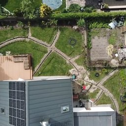 M&S Garden - Widok z góry na ogród w trakcie prac ziemnych, wykopane rowy w trawie, częściowo widoczny dom z panelami słonecznymi, plac zabaw z czerwoną zjeżdżalnią w rogu, w tle drzewa i basen.