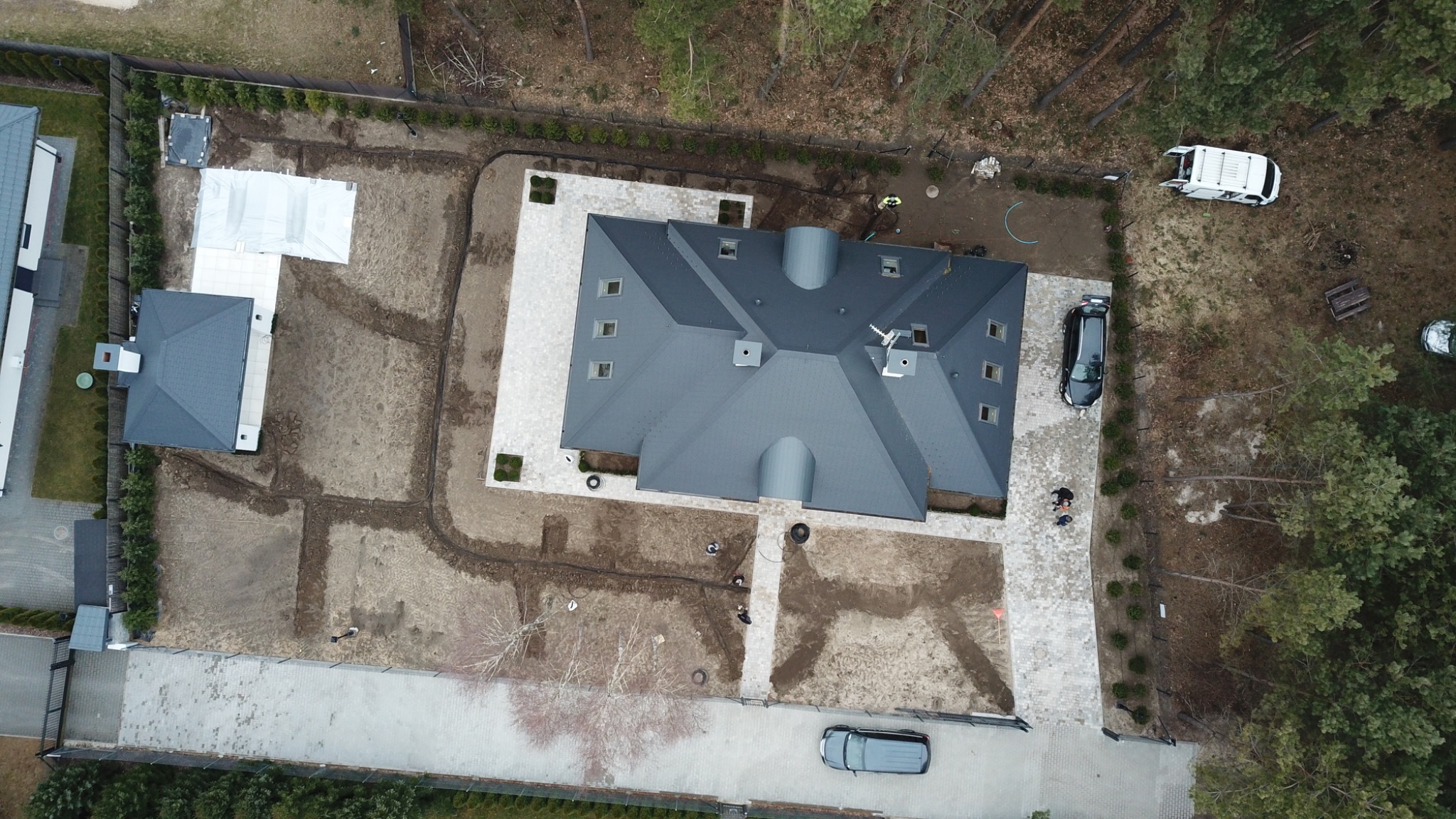 Widok z lotu ptaka na posesję w trakcie prac ogrodowych: ułożona kostka brukowa wokół domu, przygotowane rabaty, widoczne zarysy przyszłych nasadzeń, częściowo rozłożona folia na gruncie, samochody...