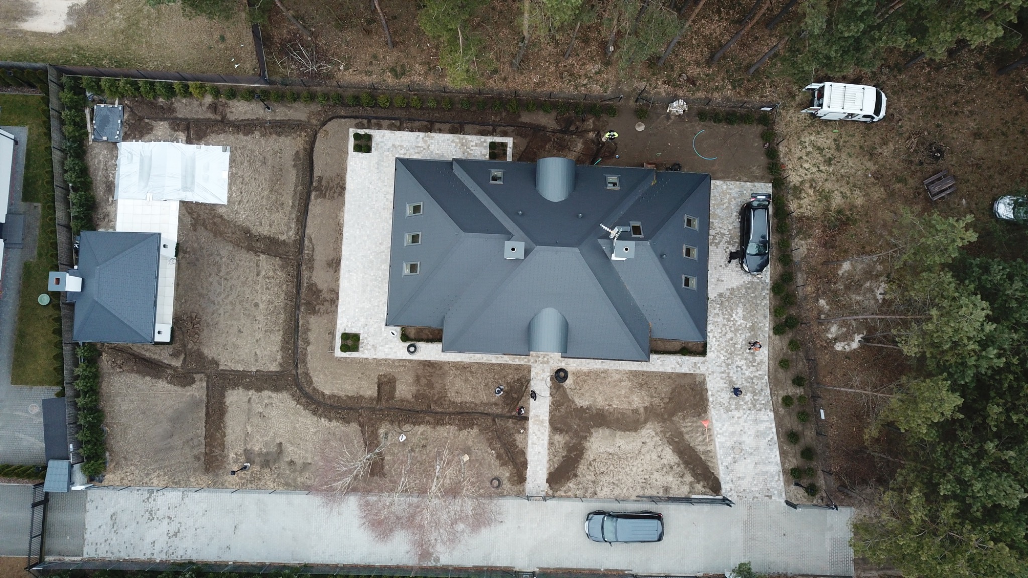 Widok z drona na posesję w Otwocku w trakcie prac ogrodowych, widoczny dom z szarym dachem, ułożona kostka brukowa, przygotowany teren pod trawnik i system nawadniania, pracownicy w żółtych...