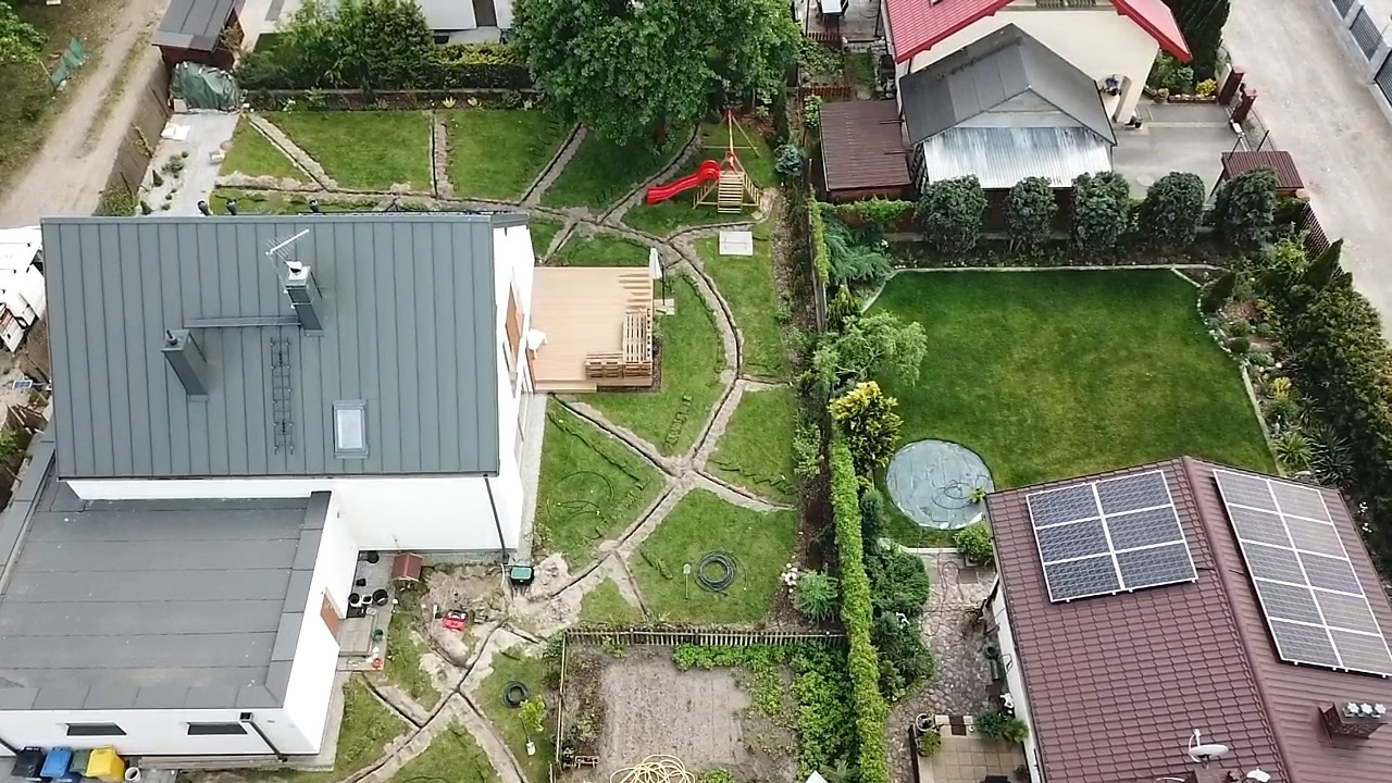 Widok z góry na ogród w trakcie instalacji systemu nawadniania, widoczne wykopy pod rury, trawnik, taras, plac zabaw i panele słoneczne na dachu sąsiedniego domu.