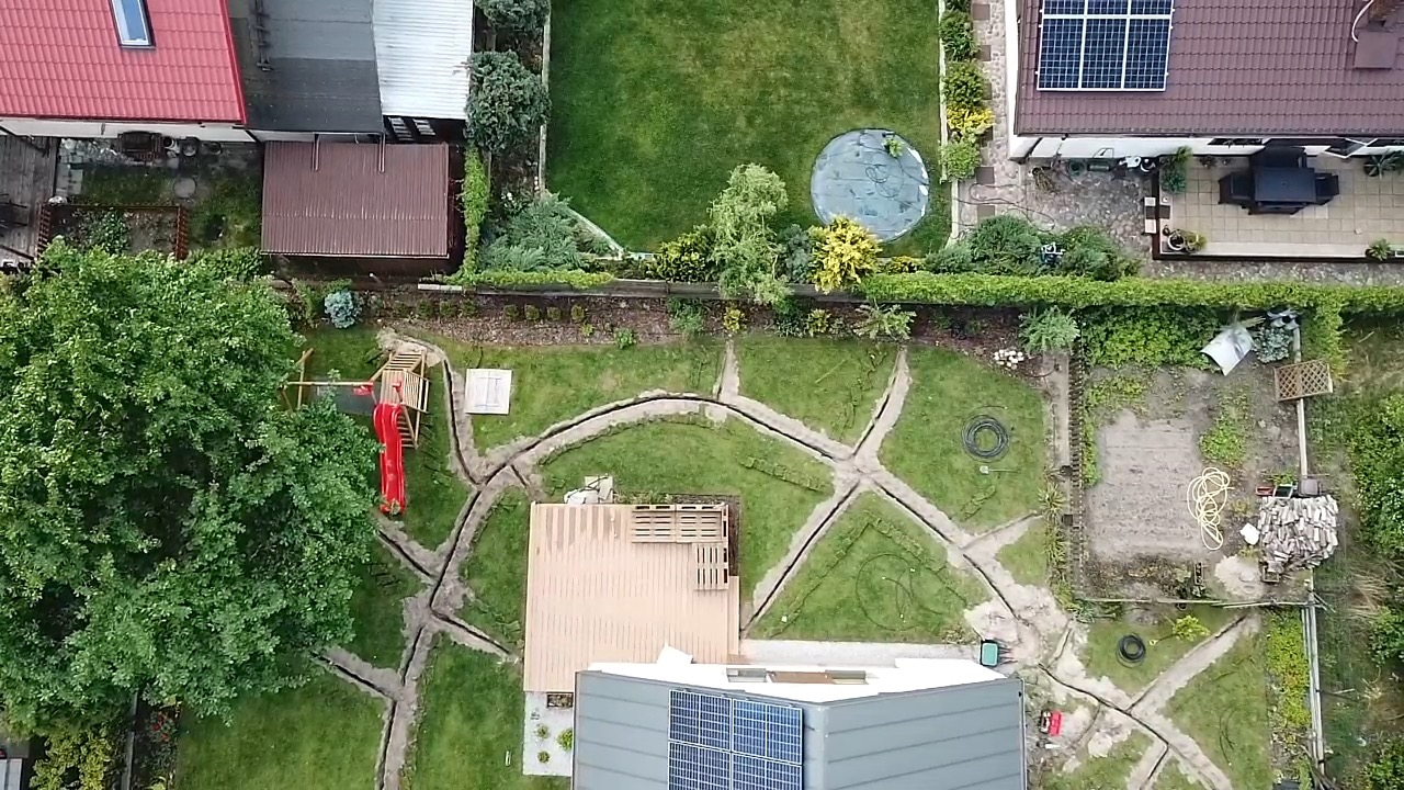 Widok z góry na ogród z wykopanymi rowami pod system nawadniania, częściowo zadaszony taras, plac zabaw dla dzieci z czerwoną zjeżdżalnią i panele słoneczne na dachu domu.