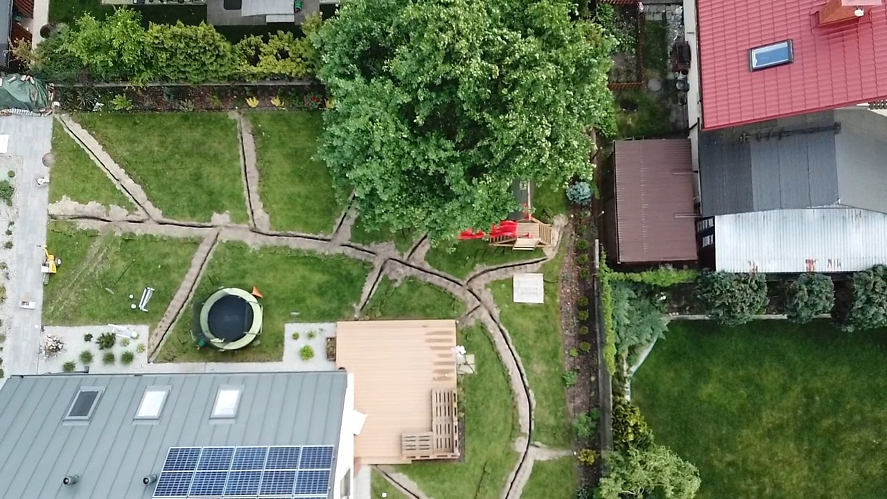 Widok z góry na ogród z wykopanymi rowami pod system nawadniania, trawnik, drzewo, trampolina i plac zabaw dla dzieci.