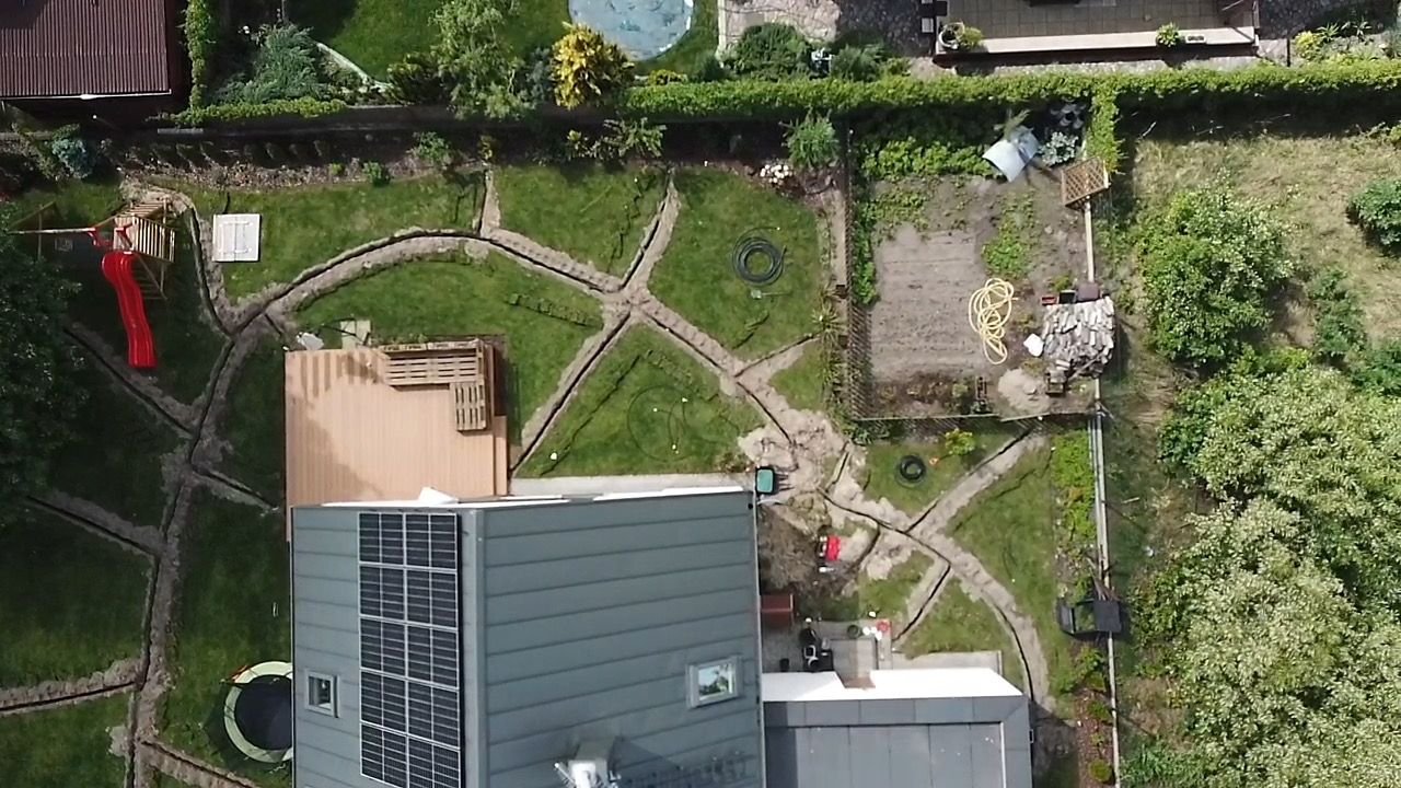 Widok z góry na ogród w trakcie prac ziemnych, wykopane rowy w trawie, częściowo widoczny dom z panelami słonecznymi, plac zabaw z czerwoną zjeżdżalnią w rogu, w tle drzewa i basen.