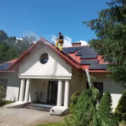 Pracownik w żółtym kombinezonie zabezpieczony linami myje dach domu z panelami słonecznymi za pomocą myjki ciśnieniowej, widoczna piana i para wodna, na tle błękitne niebo.