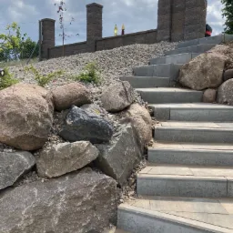 Schody z jasnoszarej kostki prowadzące w górę, otoczone dużymi, naturalnymi kamieniami i drobnym żwirem, z widocznym murem z kostki i młodym drzewkiem w tle.