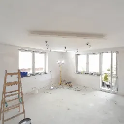 Wnętrze w trakcie prac przygotowawczych do wykończenia, widoczna drabina, lampy robocze oraz ściany po wstępnym wyrównaniu.