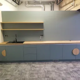 Nowoczesna kuchnia biurowa z szaro-zielonymi szafkami, drewnianym blatem i okrągłymi uchwytami, uzupełniona o dwie półki i czarny zlew.