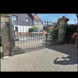 Otwarta, automatyczna brama wjazdowa z czarnego metalu z ozdobnymi elementami, zamontowana na kamiennych słupkach, z widocznym mechanizmem otwierającym, na tle brukowanego podjazdu i zabudowy...