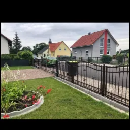 Nowoczesne, ciemne ogrodzenie metalowe z automatyczną bramą wjazdową, widok na zadbany trawnik, klomb z kwiatami i zabudowę jednorodzinną w tle.