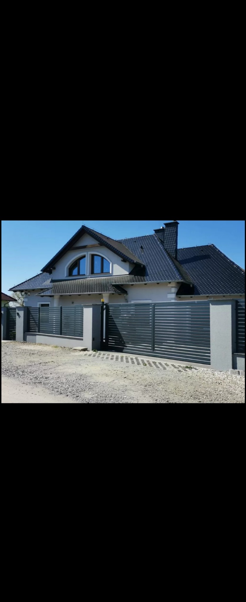 Nowoczesna, przesuwna brama wjazdowa z poziomych, metalowych profili w kolorze antracytowym, zamontowana na betonowych słupkach, na tle domu z ciemną dachówką.