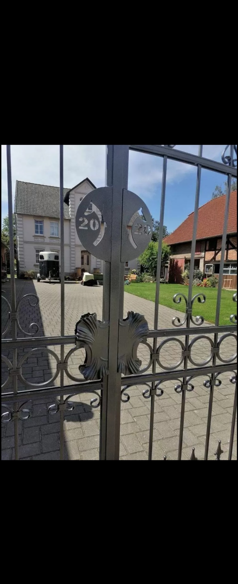 Metalowa brama wjazdowa z dekoracyjnymi elementami i numerem posesji wyciętym laserowo w metalu, na tle brukowanej drogi i budynków.