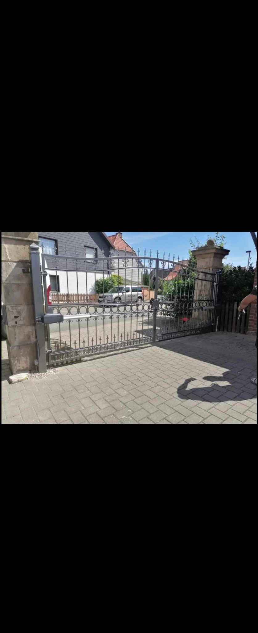 Otwarta, automatyczna brama wjazdowa z czarnego metalu z ozdobnymi elementami, zamontowana na kamiennych słupkach, z widocznym mechanizmem otwierającym, na tle brukowanego podjazdu i zabudowy...