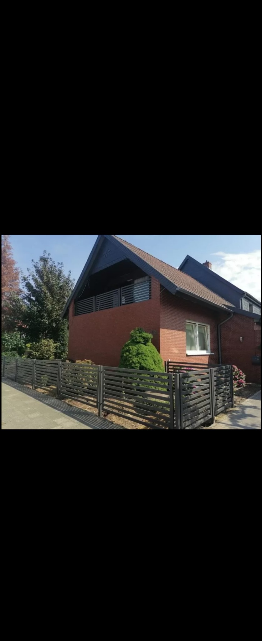 Nowoczesne, poziome ogrodzenie panelowe w kolorze antracytowym przed domem z czerwonej cegły, z widocznym balkonem z podobnymi panelami i starannie przystrzyżonym krzewem.