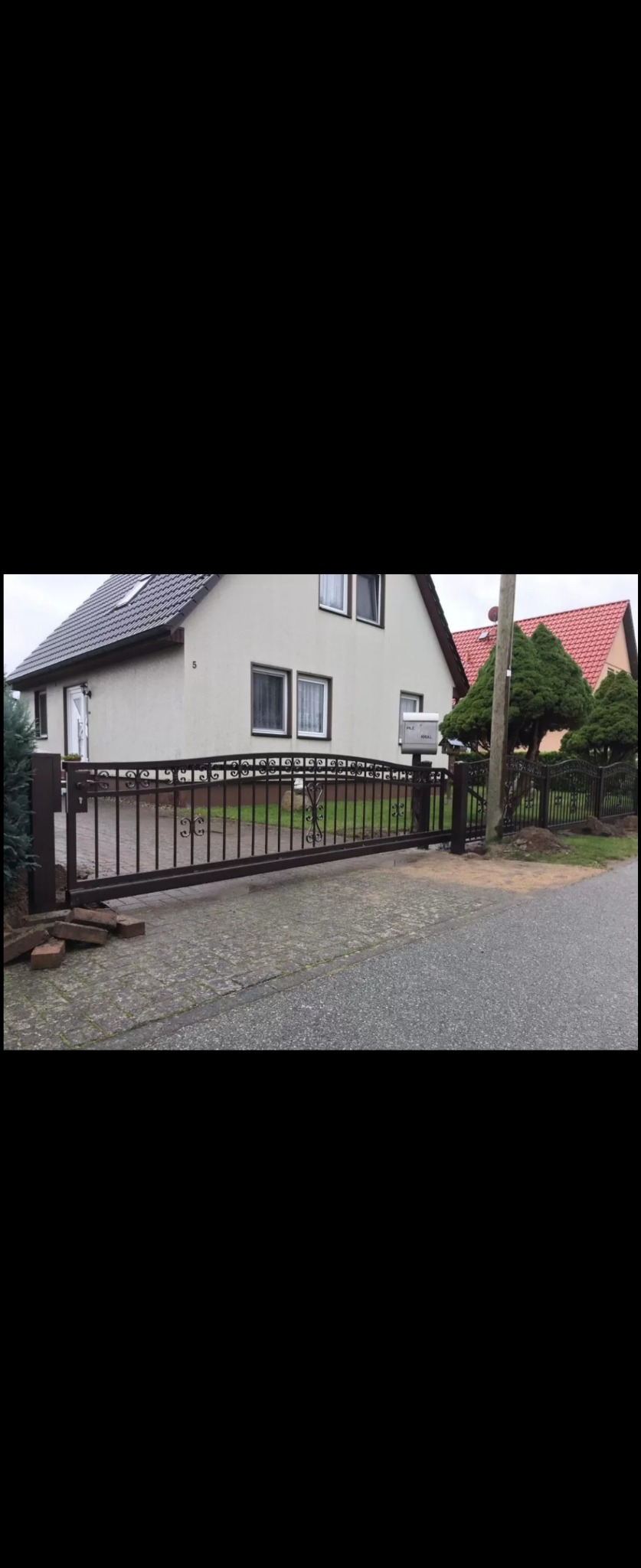Przesuwna, metalowa brama wjazdowa w kolorze ciemnego brązu, z ozdobnymi elementami, częściowo otwarta, przed domem jednorodzinnym z jasną elewacją i skrzynką pocztową.