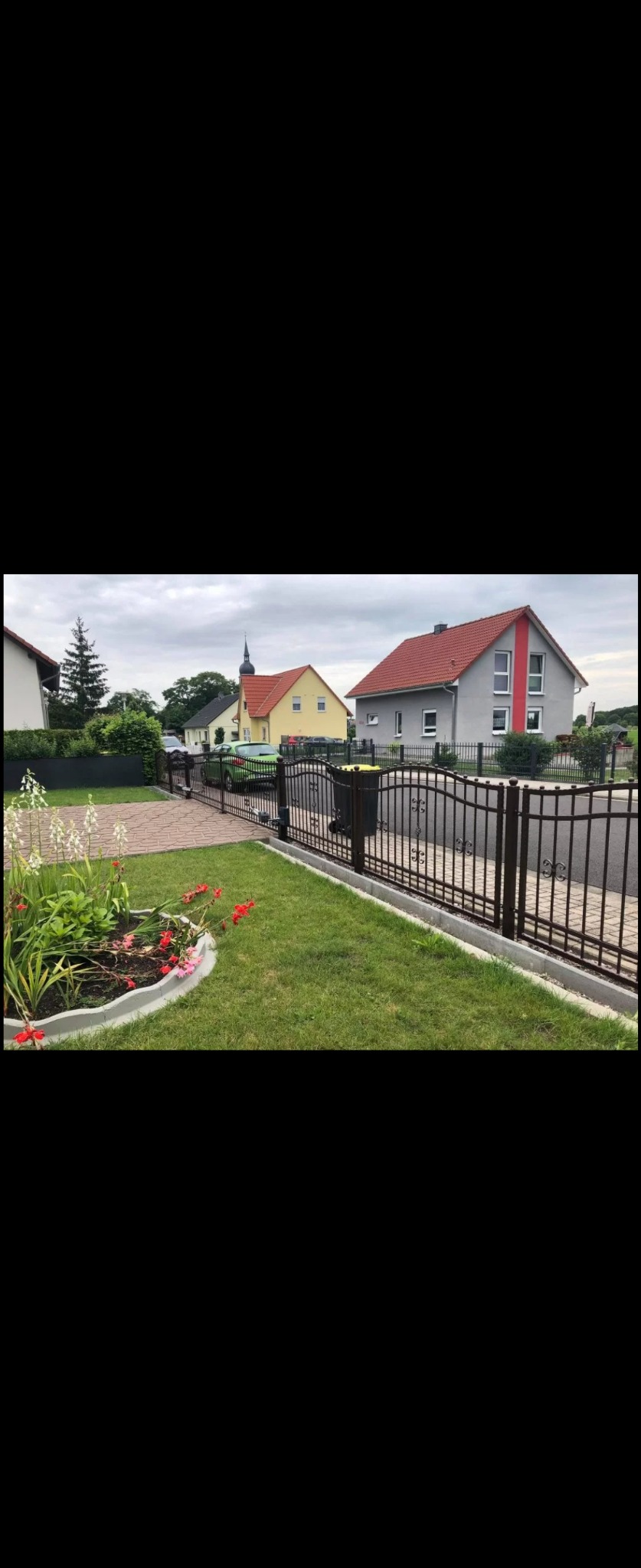 Nowoczesne, ciemne ogrodzenie metalowe z automatyczną bramą wjazdową, widok na zadbany trawnik, klomb z kwiatami i zabudowę jednorodzinną w tle.