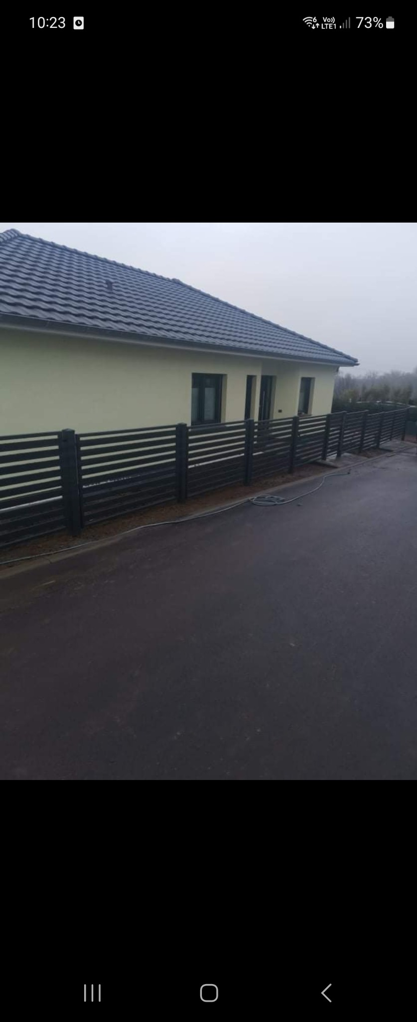 Nowoczesne ogrodzenie z poziomych, czarnych profili zamontowane przy jasnym domu z ciemną dachówką, widoczne na mokrym podjeździe w pochmurny dzień.