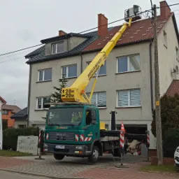 Żółty podnośnik koszowy na zielonym samochodzie ciężarowym, z operatorem w koszu, ustawiony przy dwupiętrowym budynku z czerwonym dachem; widoczne linie energetyczne nad dachem.