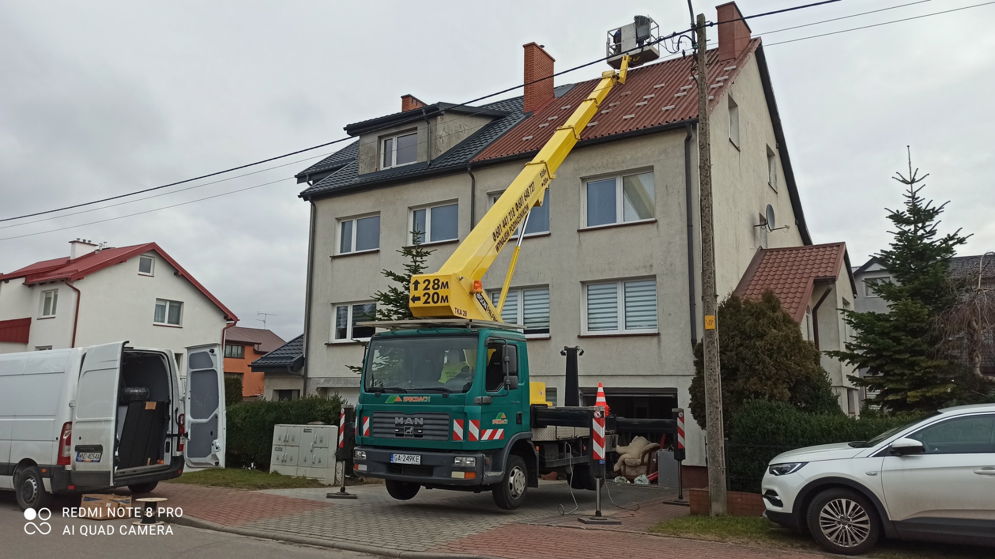 Żółty podnośnik koszowy na zielonym samochodzie ciężarowym, z operatorem w koszu, ustawiony przy dwupiętrowym budynku z czerwonym dachem; widoczne linie energetyczne nad dachem.