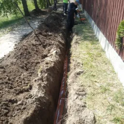 Wykopany wąski rów wzdłuż ogrodzenia z pomarańczowymi rurami w środku; robotnicy w oddali, jeden z łopatą przy rowie, trawa i drzewa w tle.