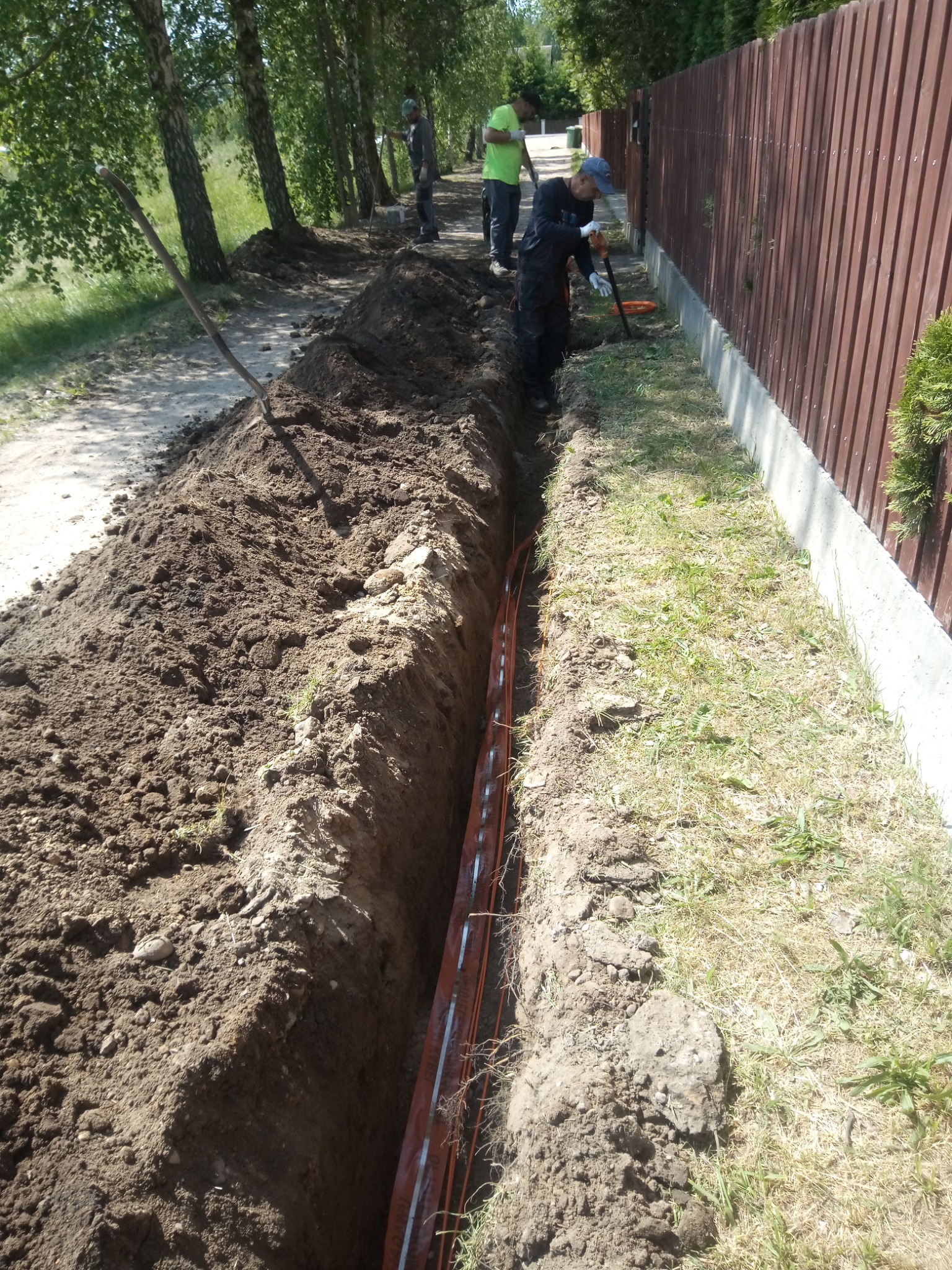 Wykopany wąski rów wzdłuż ogrodzenia z pomarańczowymi rurami w środku; robotnicy w oddali, jeden z łopatą przy rowie, trawa i drzewa w tle.