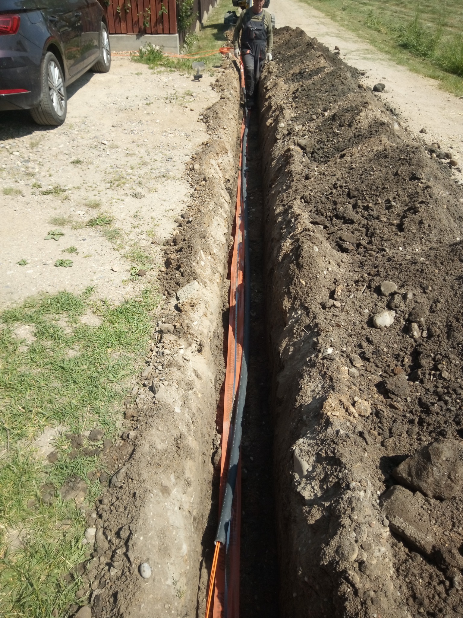 Wąski wykop w ziemi z pomarańczowymi rurami wewnątrz, pracownik w kombinezonie na końcu wykopu, samochód osobowy zaparkowany obok.