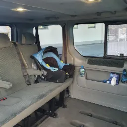 Wnętrze busa z zamontowanym fotelikiem dziecięcym na tylnej kanapie. Widoczna butelka wody i chusteczki higieniczne w kieszeni drzwi.