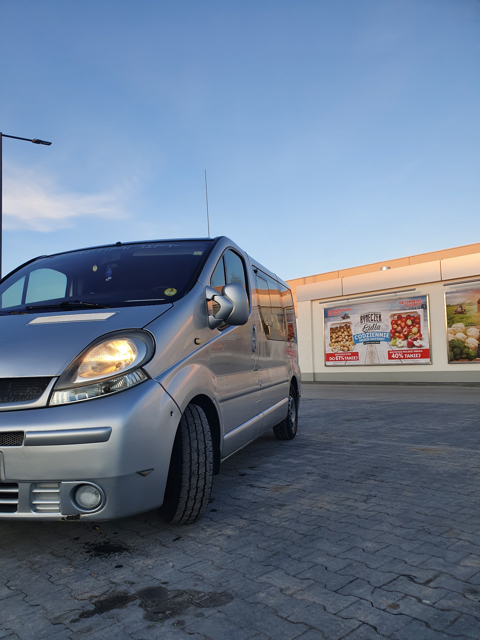Srebrny van Renault Trafic z włączonym reflektorem, zaparkowany na kostce brukowej przed budynkiem z reklamami sklepu Lidl, widok z niskiej perspektywy.