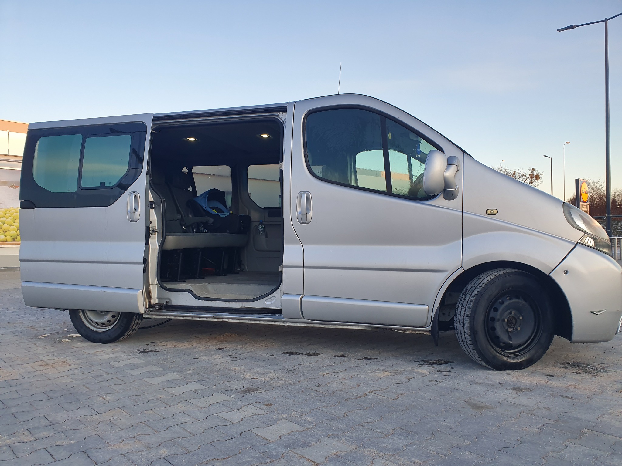 Srebrny van z otwartymi drzwiami przesuwnymi, widoczne wnętrze z fotelikiem dziecięcym, zaparkowany na szarej kostce brukowej na tle błękitnego nieba i słupów oświetleniowych.