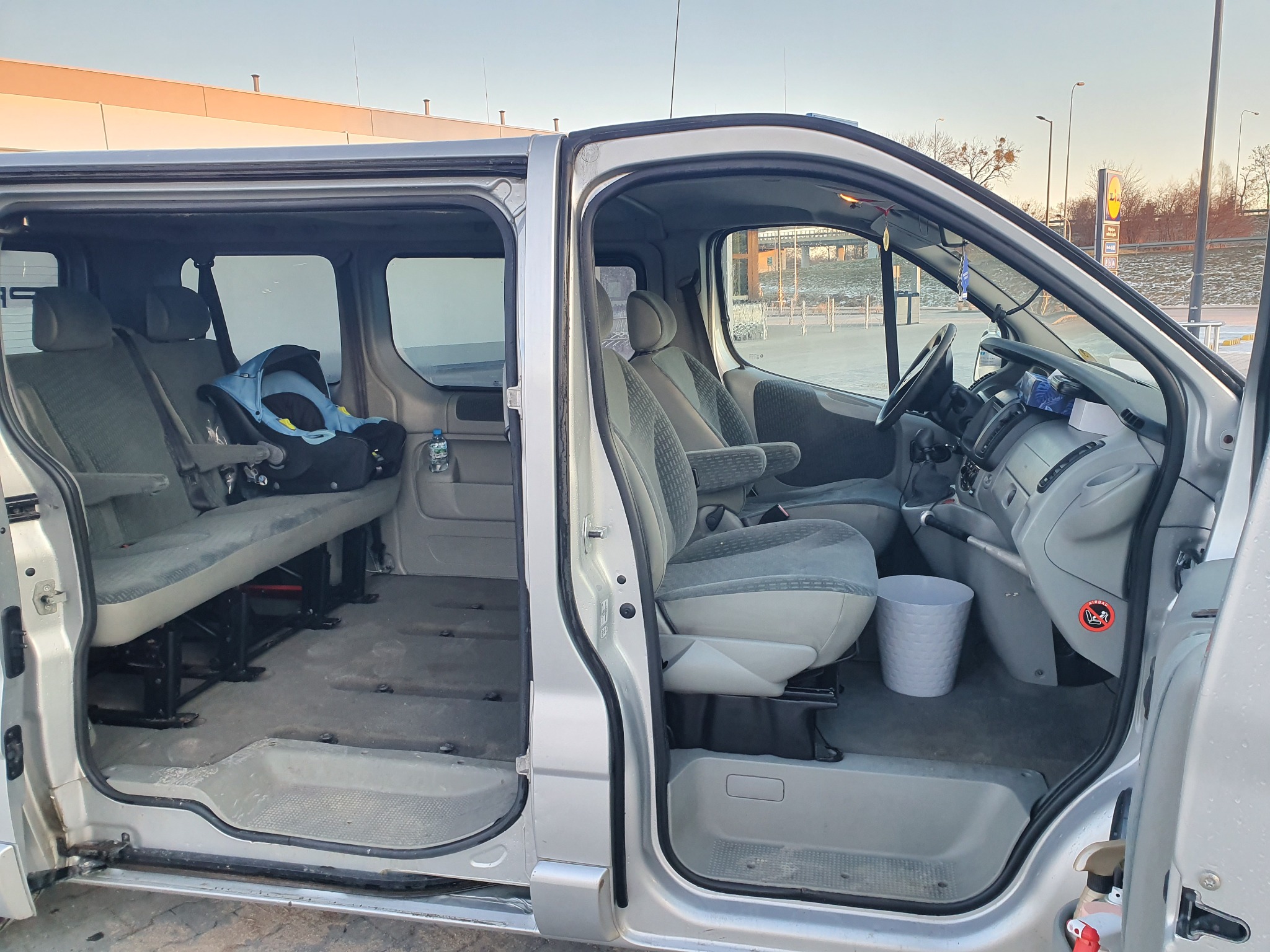 Wnętrze srebrnego vana z otwartymi drzwiami bocznymi, widoczne fotele pasażerskie, fotelik dziecięcy z tyłu, kosz na śmieci z przodu, butelka wody i naklejka ostrzegawcza na desce rozdzielczej.