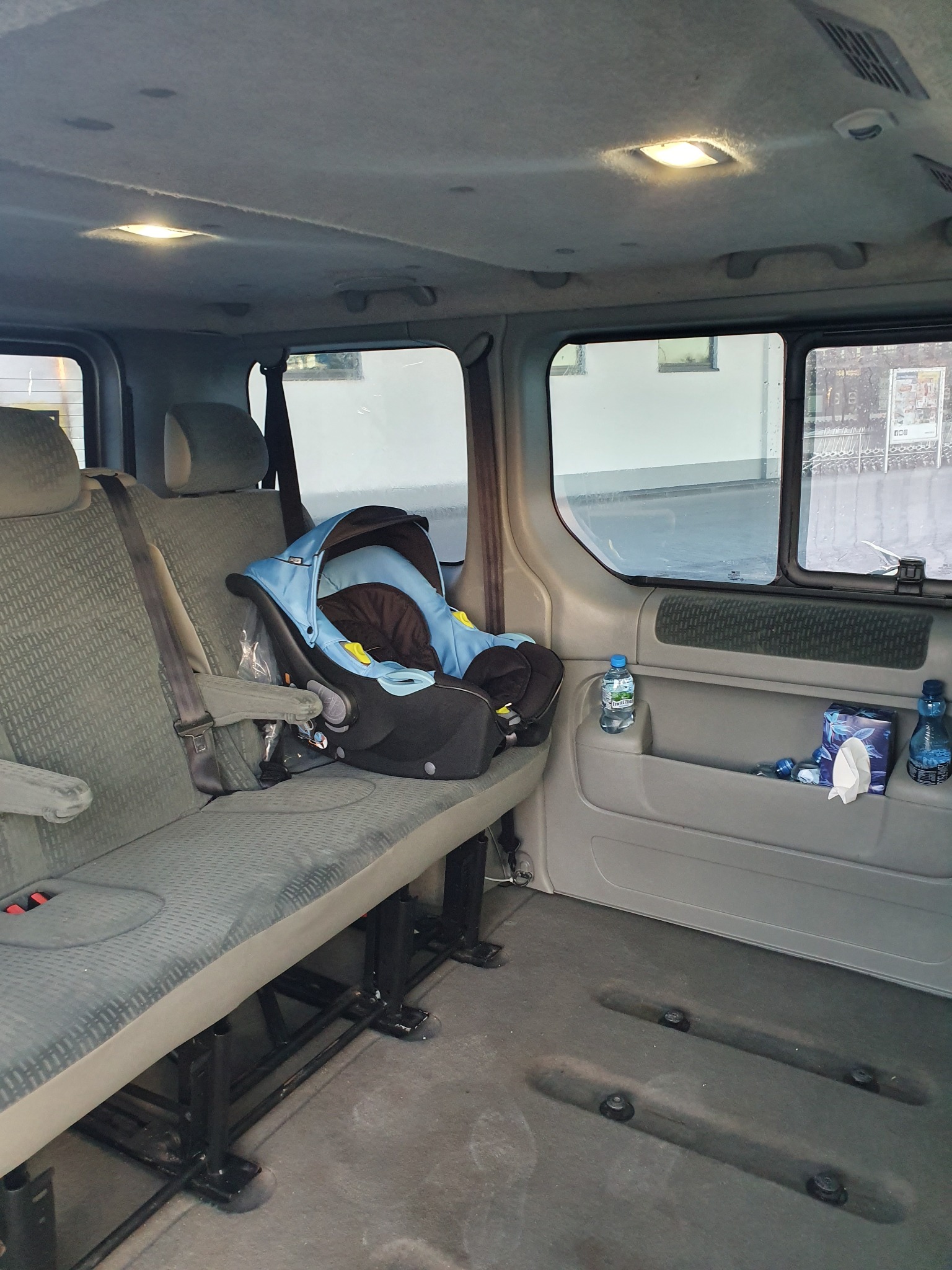 Wnętrze busa z zamontowanym fotelikiem dziecięcym na tylnej kanapie. Widoczna butelka wody i chusteczki higieniczne w kieszeni drzwi.