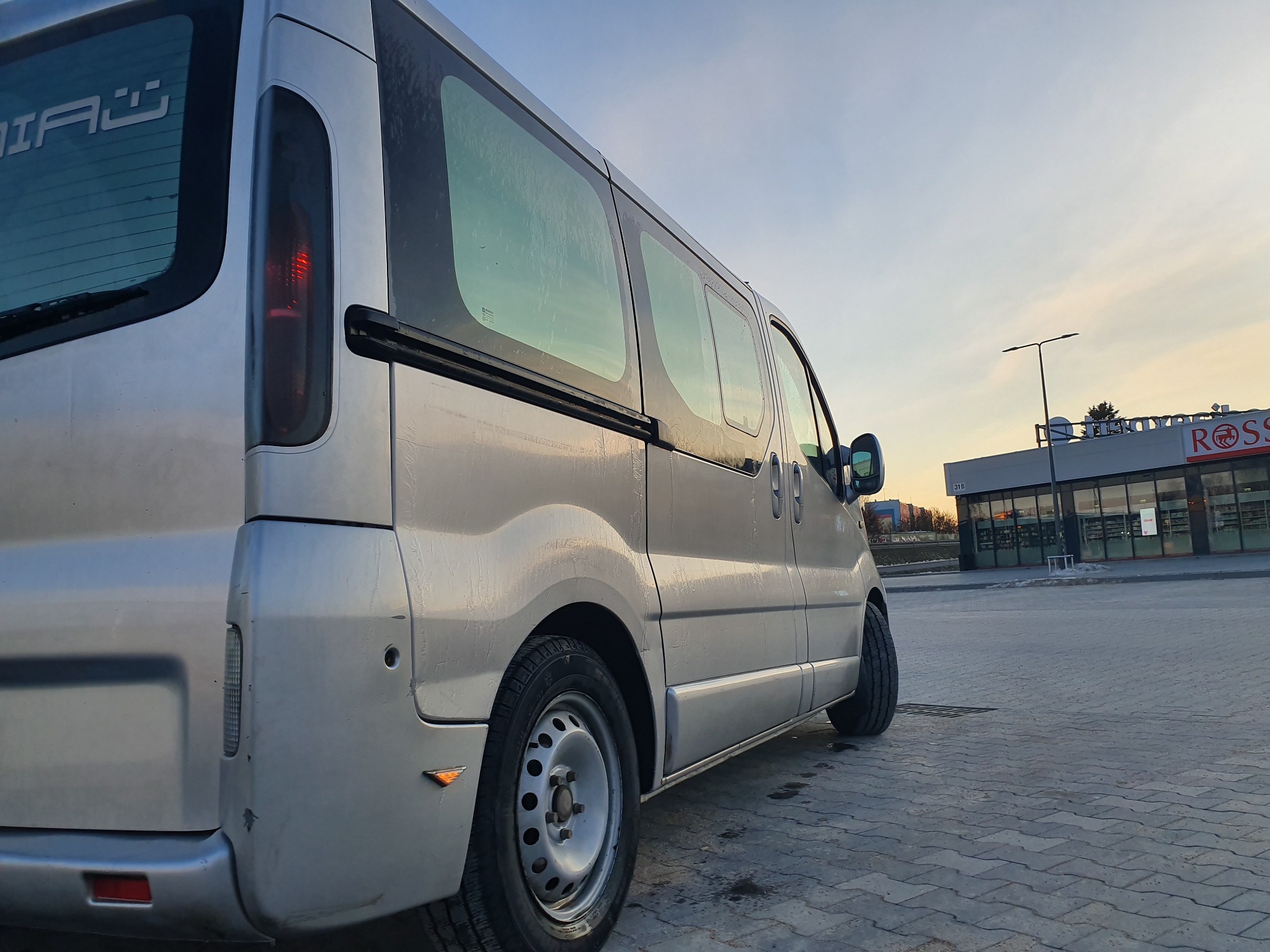 Srebrny van z przesuwnymi drzwiami zaparkowany na brukowanym parkingu, widok z tyłu i boku, z widocznym budynkiem sklepu w tle.