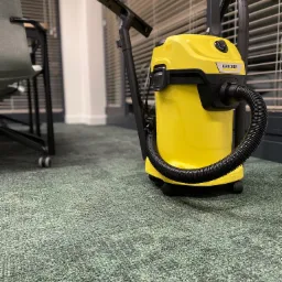 Żółty odkurzacz Karcher na zielonej wykładzinie biurowej, widoczne biurko i żaluzje w tle.