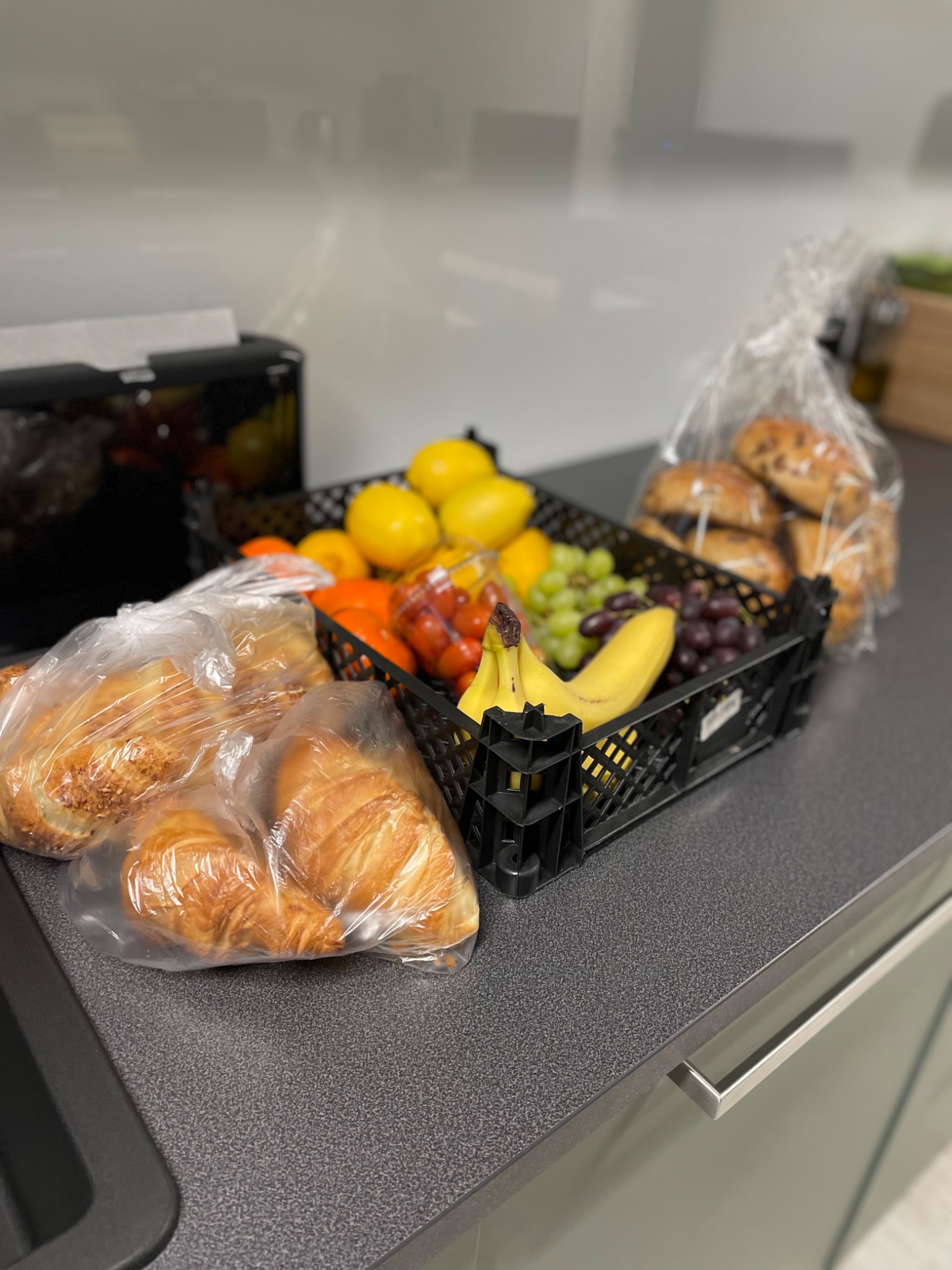 Kosz z owocami (banany, winogrona, cytryny, pomarańcze, pomidorki koktajlowe) oraz zapakowane w folię rogaliki i bułki leżące na blacie w biurowej kuchni.