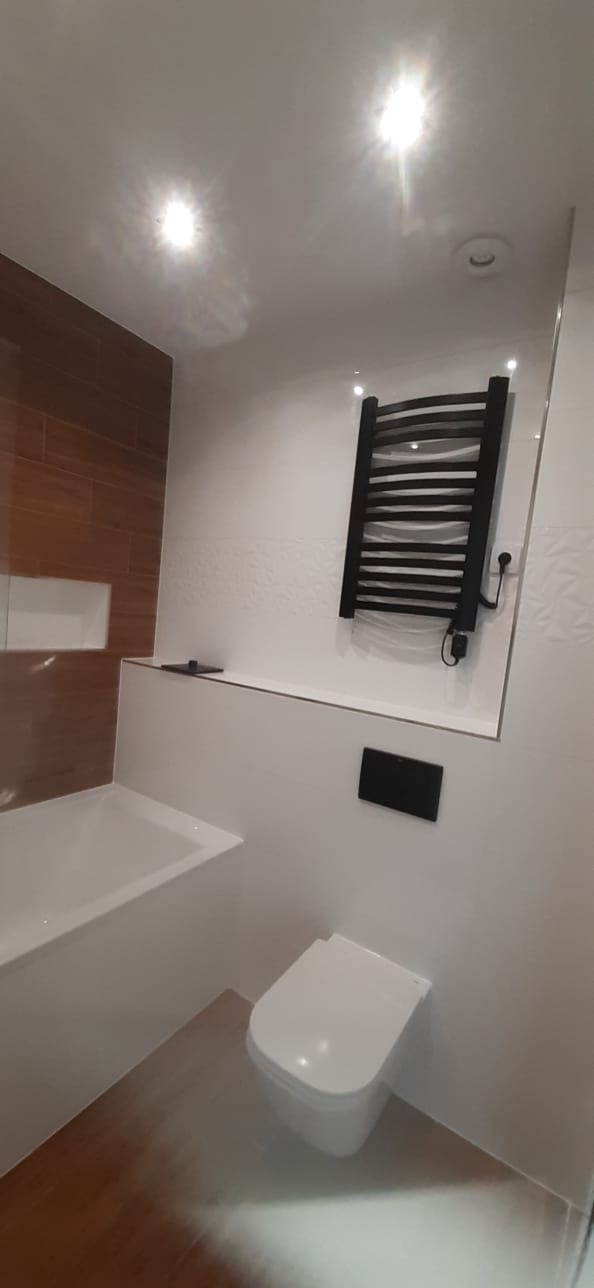 Nowoczesna łazienka z drewnopodobnymi płytkami na podłodze i części ściany, biała armatura, czarny grzejnik drabinkowy z przewodem, minimalistyczna spłuczka podtynkowa.