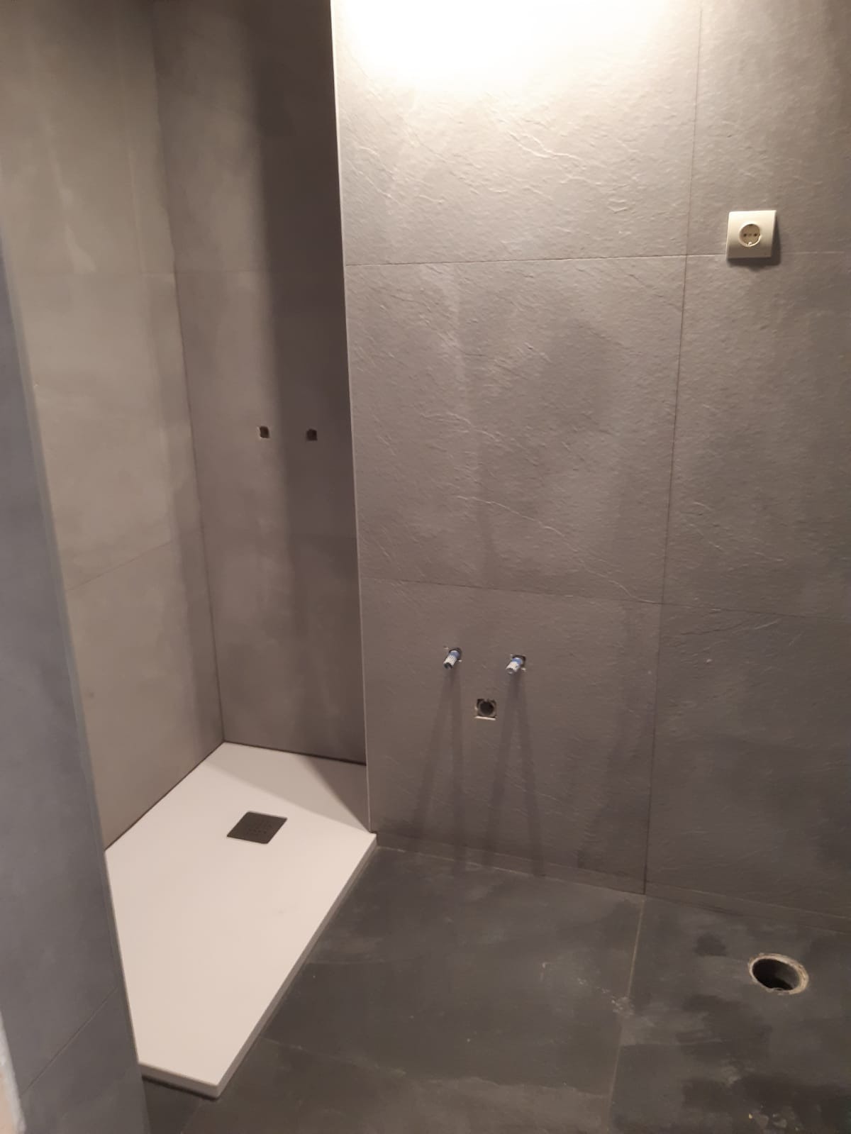 Surowa łazienka w trakcie wykończenia: szare płytki imitujące kamień, biały brodzik z odpływem kwadratowym, wystające rury instalacji wodnej i gniazdko elektryczne na ścianie.